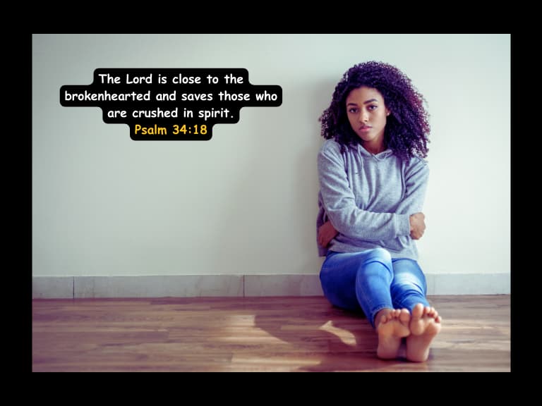 a woman sitting on the floor looking sad with her hands folded together in a hugging position with the bible verse for healing a broken heart displayed reading The Lord is close to the brokenhearted and saves those who are crushed in spirit.
Psalm 34:18