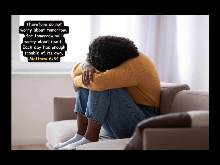 woman wearing a yellow sweater putting her head in her knees feeling anxious with the bible verse Matthew 6:34 hanging beside her